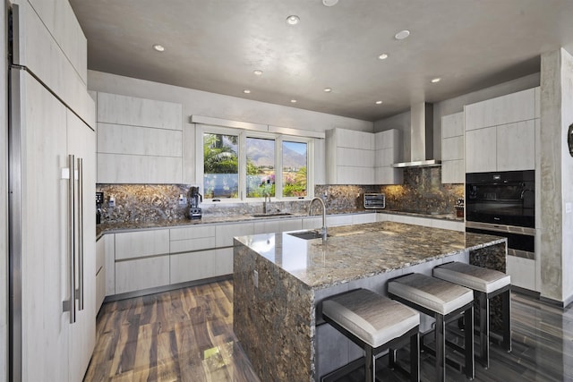 kitchen featuring dark stone countertops, a spacious island, dark wood-type flooring, a kitchen breakfast bar, and wall chimney exhaust hood