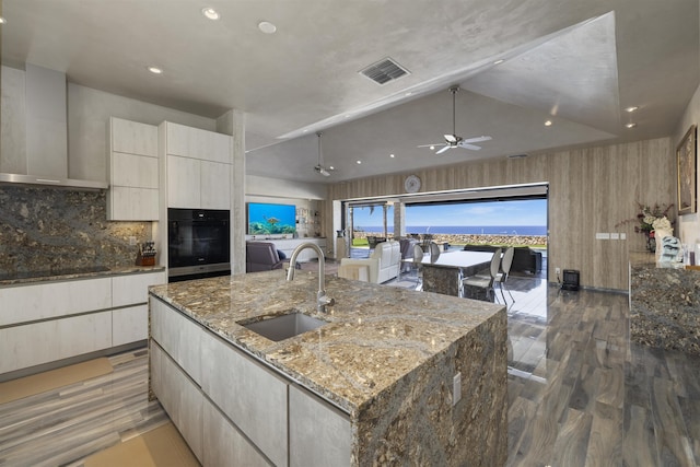 kitchen featuring a spacious island, black appliances, wall chimney range hood, sink, and light stone counters