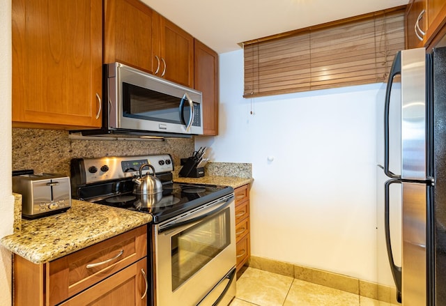 kitchen featuring light tile patterned floors, light stone counters, appliances with stainless steel finishes, decorative backsplash, and brown cabinets