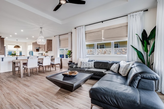 living room featuring ceiling fan with notable chandelier, a healthy amount of sunlight, a tray ceiling, and light hardwood / wood-style flooring