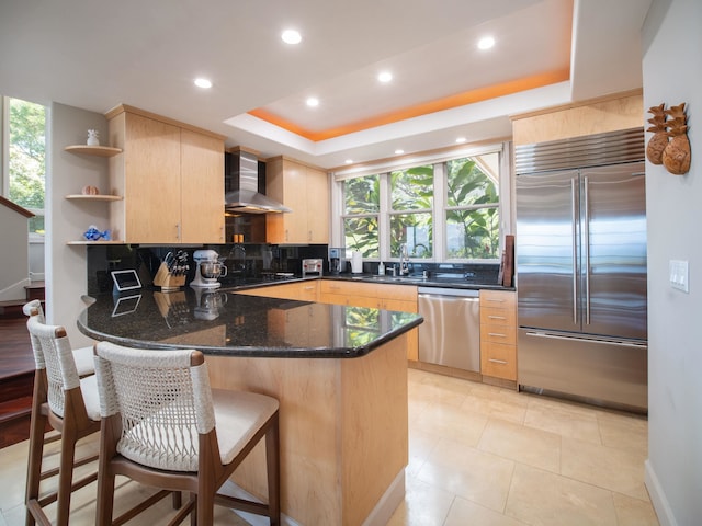 kitchen with appliances with stainless steel finishes, tasteful backsplash, wall chimney range hood, dark stone countertops, and light brown cabinetry