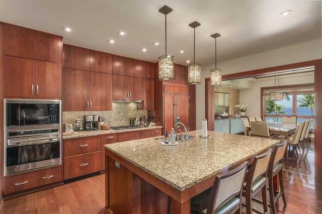 kitchen featuring sink, tasteful backsplash, built in appliances, hanging light fixtures, and an island with sink