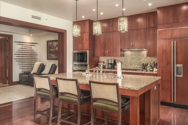 kitchen featuring built in appliances, dark wood-type flooring, tasteful backsplash, and an island with sink