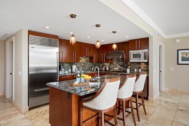 kitchen with hanging light fixtures, built in appliances, crown molding, decorative backsplash, and a kitchen island with sink