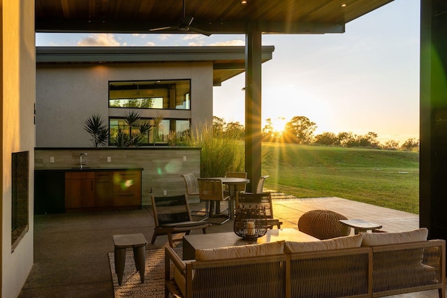 patio terrace at dusk with an outdoor kitchen, sink, a lawn, ceiling fan, and an outdoor hangout area