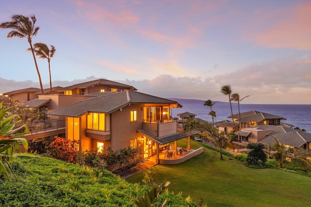 back of property at dusk with a balcony, a water view, a tiled roof, and a yard