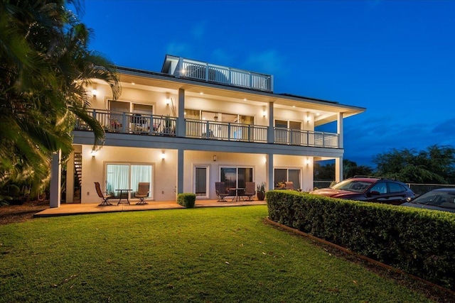 back of house at twilight featuring stucco siding, a balcony, and a lawn