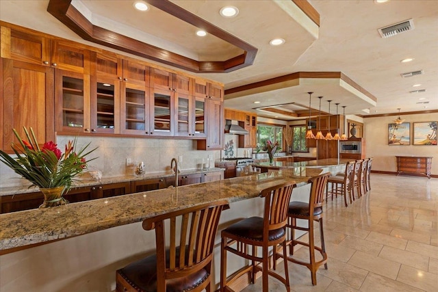 85 Lau Awa Pl, Lahaina, HI 96761 - kitchen featuring wall chimney exhaust hood, a tray ceiling, stone tile flooring, ornamental molding, and a breakfast bar