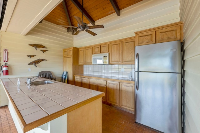 kitchen with tile counters, stainless steel refrigerator, ceiling fan, beam ceiling, and sink