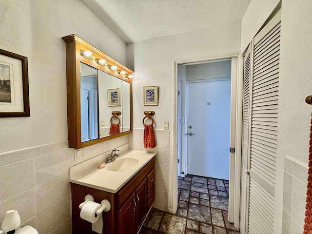 bathroom featuring stone finish flooring, tile walls, a wainscoted wall, vanity, and a textured ceiling