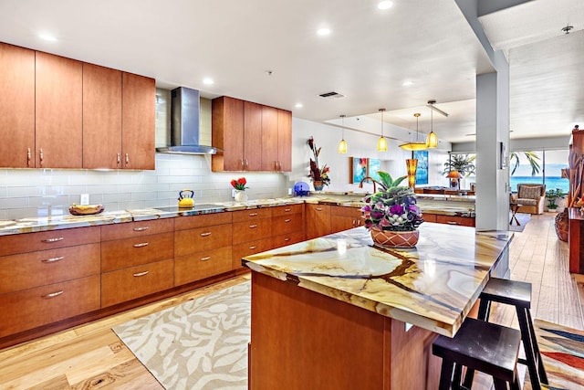 kitchen featuring wall chimney exhaust hood, light hardwood / wood-style floors, a kitchen bar, kitchen peninsula, and light stone counters