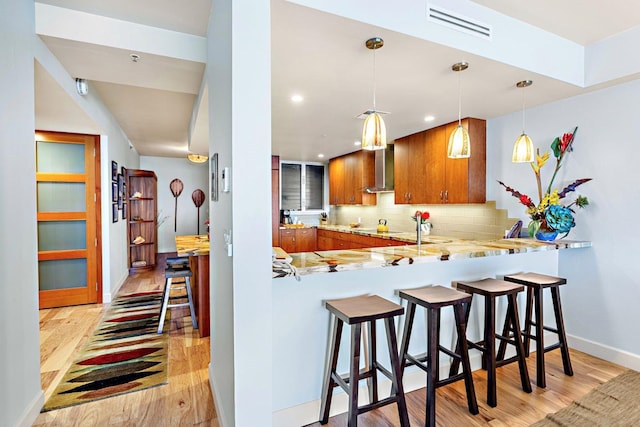 kitchen with hanging light fixtures, light wood-type flooring, wall chimney range hood, kitchen peninsula, and decorative backsplash