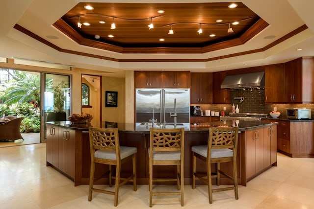 kitchen featuring a raised ceiling, wall chimney range hood, built in refrigerator, tasteful backsplash, and a large island