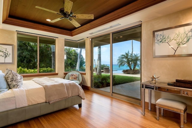 bedroom featuring access to outside, a water view, ceiling fan, a tray ceiling, and light hardwood / wood-style floors