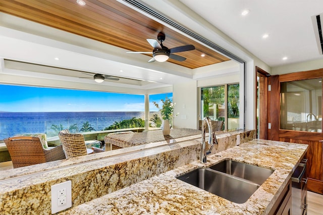 kitchen featuring light stone counters, wood ceiling, ceiling fan, sink, and a water view