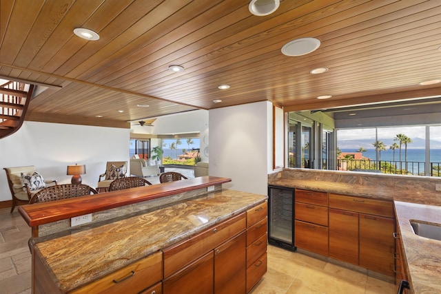 bar with stone counters, ceiling fan, beverage cooler, a water view, and wood ceiling