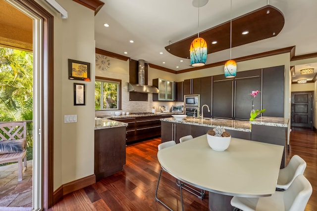 kitchen featuring wall chimney exhaust hood, dark brown cabinetry, decorative light fixtures, dark wood-type flooring, and light stone counters