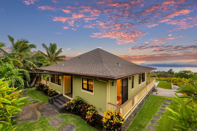 back of house at dusk featuring roof with shingles, a lawn, and covered porch