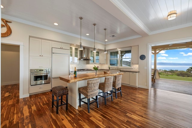 kitchen with paneled refrigerator, wall chimney exhaust hood, glass insert cabinets, stainless steel oven, and a breakfast bar area