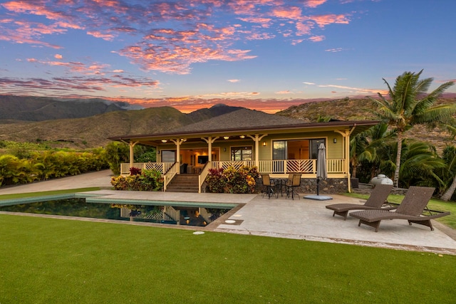 back of house at dusk with a patio, an outdoor pool, a mountain view, and a lawn