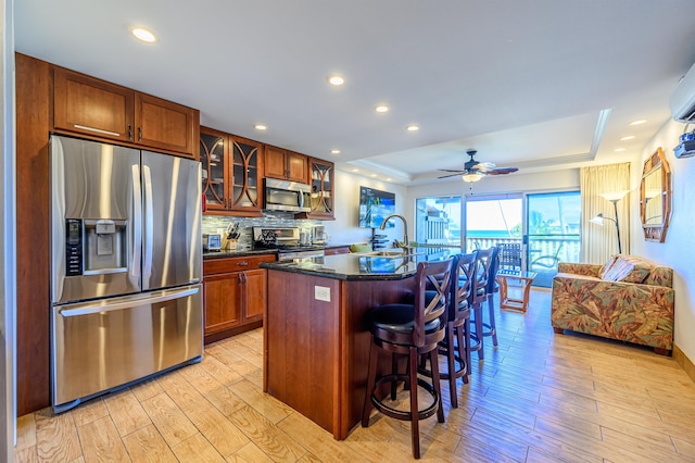 kitchen with backsplash, a tray ceiling, a center island with sink, light hardwood / wood-style floors, and stainless steel appliances