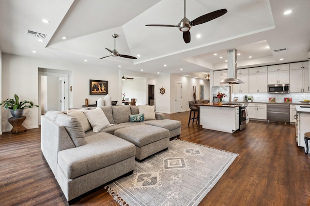 living room with a tray ceiling, visible vents, dark wood finished floors, and a ceiling fan