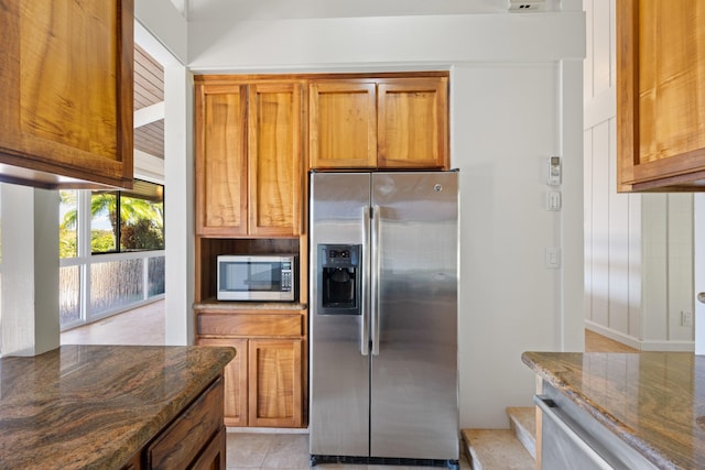 kitchen with light tile patterned floors, dark stone counters, brown cabinets, and appliances with stainless steel finishes