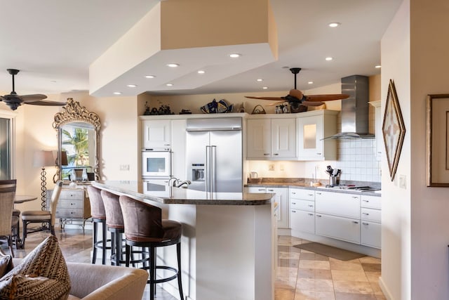 kitchen with an island with sink, white cabinetry, wall chimney range hood, decorative backsplash, and appliances with stainless steel finishes