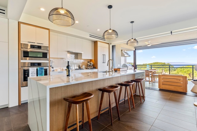 kitchen with white cabinets, stainless steel double oven, decorative light fixtures, and a center island with sink