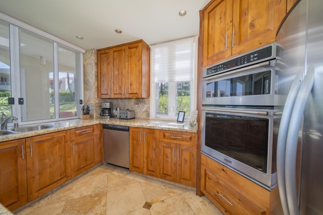 kitchen with tasteful backsplash, appliances with stainless steel finishes, brown cabinets, light stone counters, and a sink