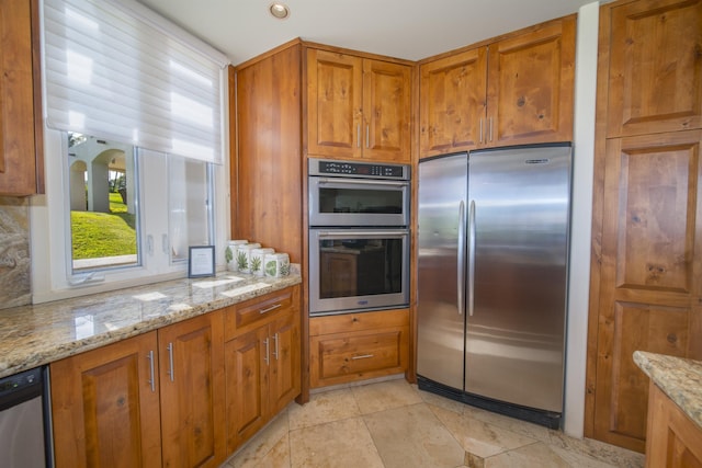 kitchen featuring light tile patterned floors, appliances with stainless steel finishes, brown cabinetry, and light stone countertops