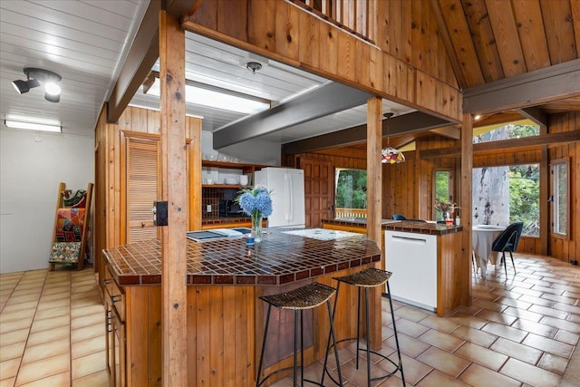 kitchen with lofted ceiling with beams, wooden walls, tile counters, kitchen peninsula, and white appliances