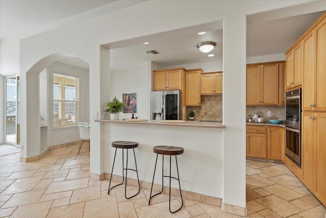 kitchen featuring light brown cabinets, a breakfast bar area, decorative backsplash, kitchen peninsula, and stainless steel appliances