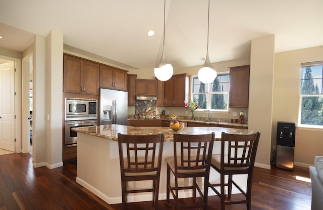 kitchen with light stone counters, a center island, stainless steel appliances, hanging light fixtures, and under cabinet range hood