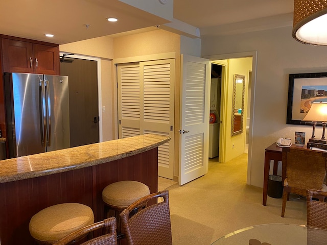 kitchen with kitchen peninsula, a kitchen bar, stainless steel refrigerator, stacked washer and dryer, and light colored carpet