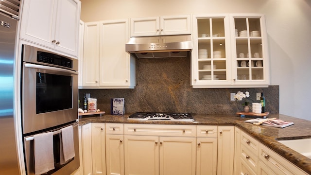 kitchen featuring under cabinet range hood, appliances with stainless steel finishes, decorative backsplash, dark stone counters, and glass insert cabinets