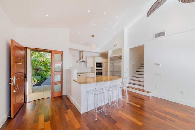 kitchen with a breakfast bar, dark wood-type flooring, wall chimney range hood, built in appliances, and high vaulted ceiling