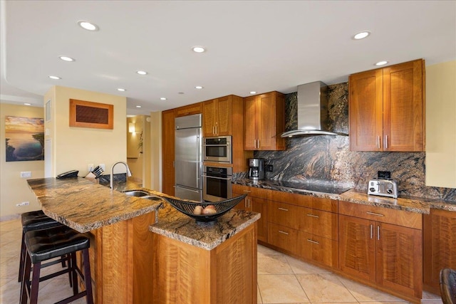 kitchen with sink, wall chimney range hood, dark stone countertops, a breakfast bar area, and appliances with stainless steel finishes