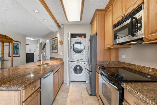 kitchen featuring stainless steel appliances, a sink, stacked washer / drying machine, dark stone countertops, and a peninsula