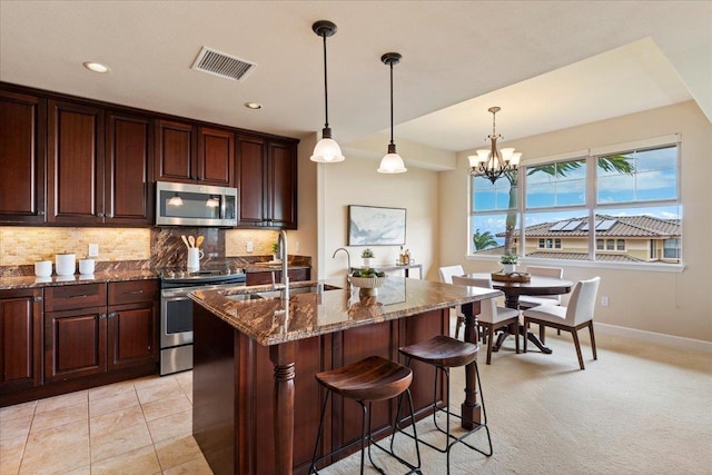 kitchen featuring appliances with stainless steel finishes, dark stone counters, an island with sink, sink, and a notable chandelier