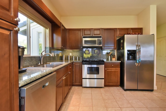 kitchen featuring stainless steel appliances, backsplash, brown cabinetry, a sink, and light stone countertops