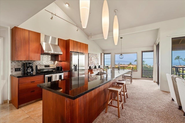 kitchen with brown cabinetry, lofted ceiling, a kitchen bar, wall chimney exhaust hood, and appliances with stainless steel finishes