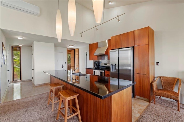 kitchen with brown cabinetry, a wall mounted air conditioner, wall chimney exhaust hood, a sink, and appliances with stainless steel finishes