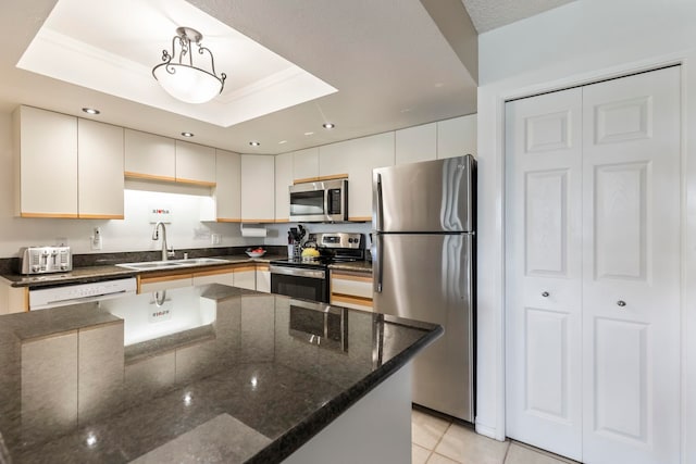 kitchen with a sink, stainless steel appliances, dark stone counters, light tile patterned flooring, and a tray ceiling