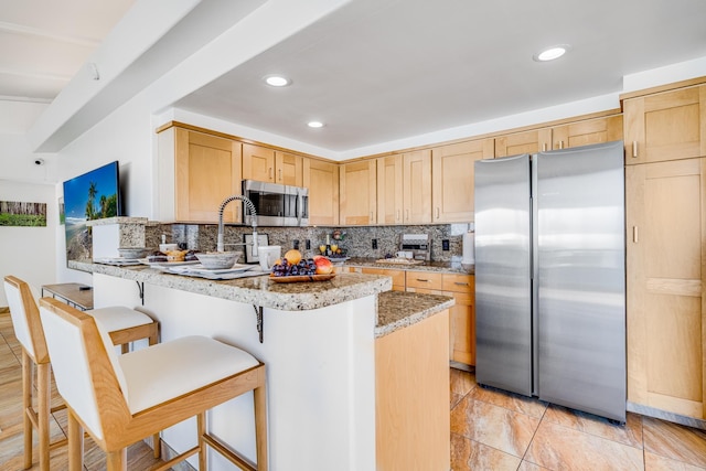 kitchen featuring a kitchen bar, light brown cabinetry, light stone counters, stainless steel appliances, and decorative backsplash