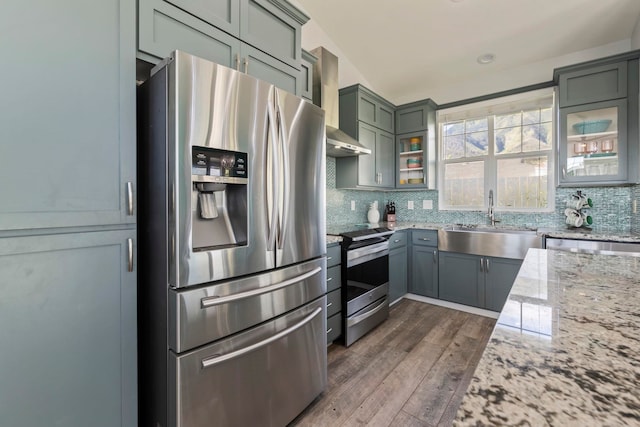 kitchen with light stone counters, stainless steel appliances, a sink, wall chimney exhaust hood, and glass insert cabinets