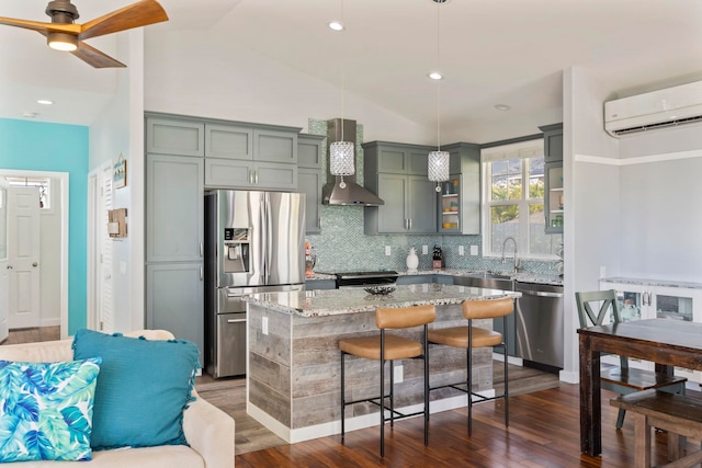 kitchen featuring a wall unit AC, stainless steel appliances, hanging light fixtures, a kitchen island, and wall chimney range hood