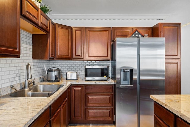 kitchen with stainless steel appliances, backsplash, brown cabinetry, a sink, and light stone countertops