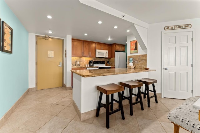 kitchen featuring white appliances, brown cabinetry, light stone counters, a breakfast bar, and a peninsula