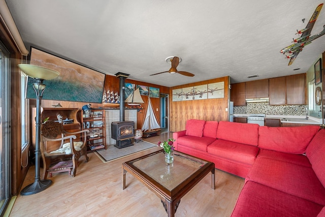 living room featuring a textured ceiling, light wood-style flooring, wood walls, a ceiling fan, and a wood stove